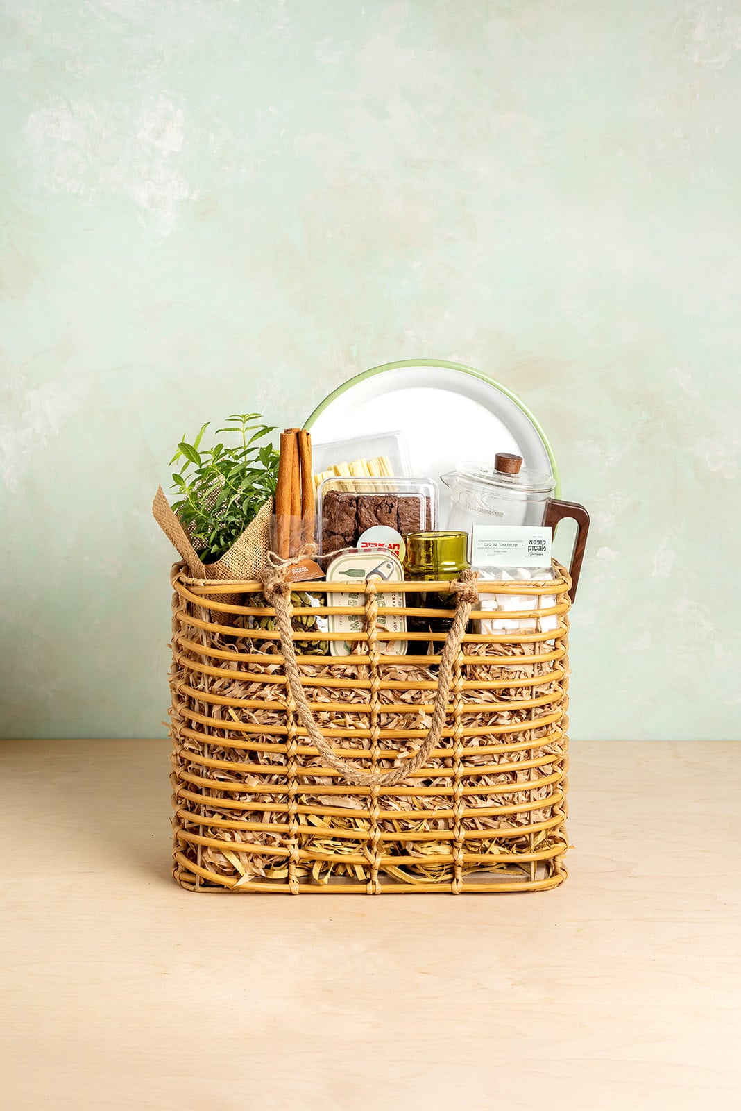 מארז פסח "החיים הטובים" של קופסא מהשוק בתוך סל נצרים עמוק, הכולל עציץ תבלין רענן, קומקום תה מזכוכית, מגש אמייל, עוגיות בראוניז, מקלות קינמון וחליטות בוטיק.